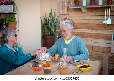 A Couple Of Attractive People, A Senior Man And Woman Sharing A Moment Of Food And Drink On A Wooden Table. Laughing They Eat Popcorn And Drink A Glass Of Beer. Two People