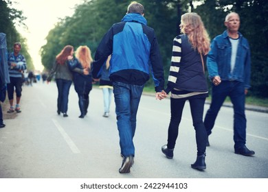 Couple, back and holding hands while walking among trees and crowds along road in New Zealand. Rear view of man, woman and people for travel, date and bonding in busy woods, park and highway