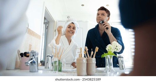 A couple in bathrobes enjoys their morning routine. The woman uses a lotion while the man shaves with an electric razor. They stand by the sink in a bright, modern bathroom, smiling and relaxed. - Powered by Shutterstock