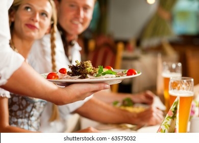 Couple In Bavarian Tracht Drinking Wheat Beer In A Typical Pub, The Waitress Is Serving The Drinks