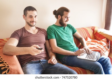 A Couple Of Bearded Brothers Having Fun In A Couch With One Of Them Typing On A Laptop Computer And The Other Holding A Tv Remote Control To Browse In His Smart Tv.