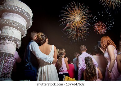 Couple Of Beautiful Stylish Newlyweds Looking At Fireworks Exploding In The Night Sky On Their Wedding Day. Behind The Wedding Cake