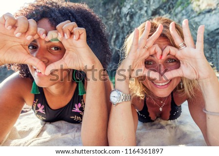 couple of beautiful women of different ethnicity showing heart shape with hands on beach vacation