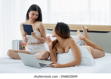 Couple Of Beautiful Young Asian Sexy Girls On White Bed Funny Talk And Laugh To Each Other While Enjoying Their Social Media From Tablet And Laptop In Bright Morning.