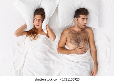 Couple In Bed. Depressed Young Woman Covers Ears With Pillow, Cant Sleep Because Of Mans Snoring, Has Sleepless Night, Lies Irritated In Bed. Family, Bed Time, Insomnia, Sleep Problems Concept