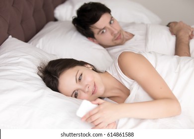 Couple in bed, happy smiling woman turned her back to man, reading message on phone from her lover, worried boyfriend lying next to her, trying to peek at screen. Cheating and infidelity concept