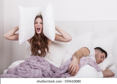 couple in bed man sleeps and snores while woman is unable to sleep closing her ears with big pillows