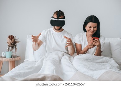 Couple in bed with man in VR headset gesturing and woman using smartphone, amused - Powered by Shutterstock