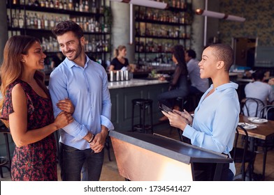 Couple Being Greeted By Maitre D Using Digital Tablet As They Arrive At Restaurant
