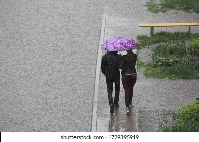 A couple in black jackets under one bright umbrella walk along the sidewalk near a large puddle with text space against the background of a bench on a spring day