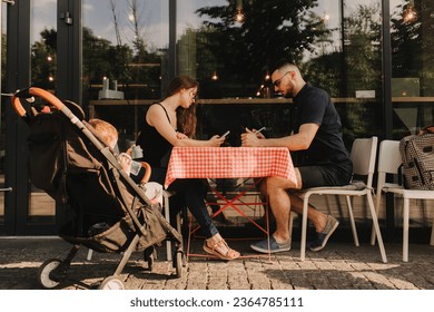 Couple are busy working on gadgets, little son is waiting in a stroller in cafe. Parents ignoring communication using devices. Freelancing and networking concept. - Shutterstock ID 2364785111