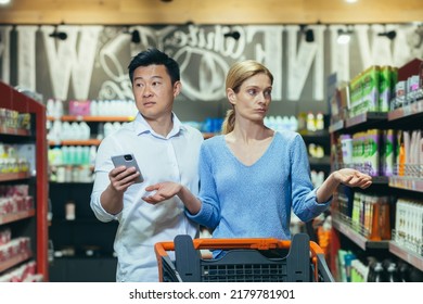 A couple of buyers, a man and a woman, confused in the supermarket, cannot choose the necessary product, an Asian man with a phone