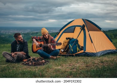 Couple Of Campers Playing Guitar While Sitting Near The Camp Fire And A Tent While Camping In The Nature