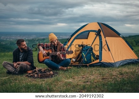 Couple of campers playing guitar while sitting near the camp fire and a tent while camping in the nature