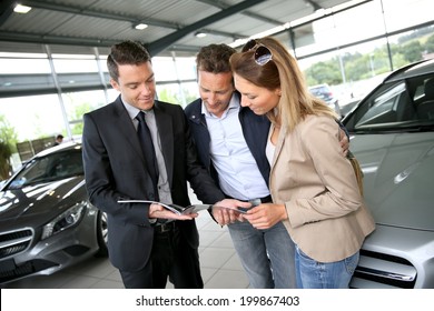 Couple In Car Dealership Looking At Brochure With Salesman