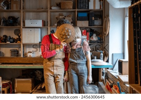 Couple of carpenters kissing at workplace. Husband boyfriend man and wife girlfriend woman fooling around hiding behind wooden product holding hands. Romance love expression affection tenderness.