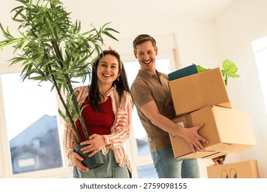 A couple carries plants and boxes while moving into their new home. They stand inside a bright room with large windows, smiling at each other as they work together.