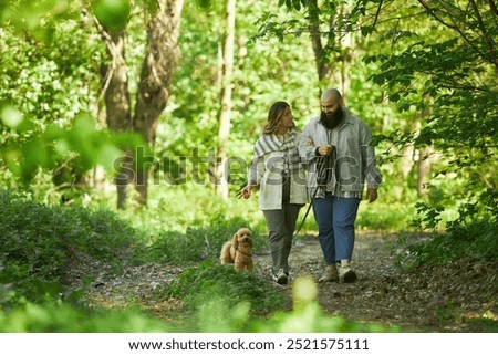 Couple casually walking their dog through vibrant green forest, enjoying nature. Both individuals smile while holding hands and leash