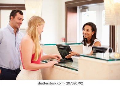 Couple Checking In At Hotel Reception Using Digital Tablet