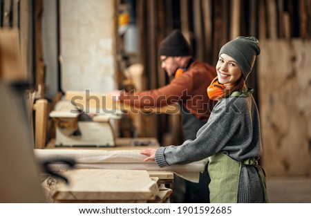 Couple of cheerful carpenters with wooden details in hands working together near workbench in professional workroom