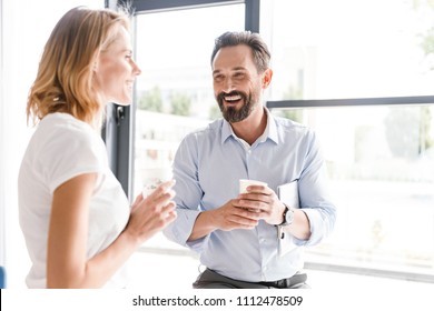 Couple Of Cheerful Colleagues Talking While Standing At The Office Window With Cups Of Coffee