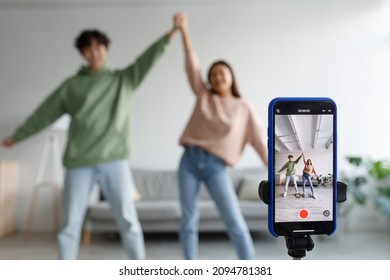 Couple of cheery Asian influencers shooting dance video for social network on cellphone, having fun at home, selective focus. Millennial bloggers streaming content for their vlog, broadcasting online