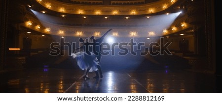 Couple of classical ballet dancers practice on theatre stage before performance. Man in training suit lifts graceful ballerina. Dance choreography rehearsal. Illuminated theatrical hall. Slow motion.
