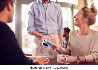 Couple In Coffee Shop Making Contactless Payment With Card For Drinks To Male Waiter