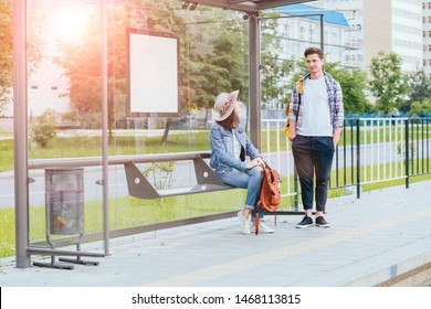 Couple communicating while waiting at bus stop. Hansome man and elegant woman with backpacks meets and talks at tram stop, outdoor. Students waiting for tram. Sun glare effect.