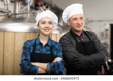 Couple of confident successful brewers, man and woman in black aprons standing with crossed arms in craft brewery against background of fermentation tanks - Powered by Shutterstock