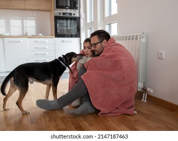 Couple Covered With Blanket Sitting On The Floor Against Radiator Trying To Warm Up While Dog Tries To Snuggle Between Them.