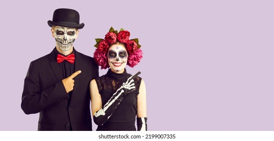 Couple in creative costumes pointing finger at copy space recommending something Halloween themed. Smiling woman and man with skull makeup together advertise product on light lilac background. - Shutterstock ID 2199007335