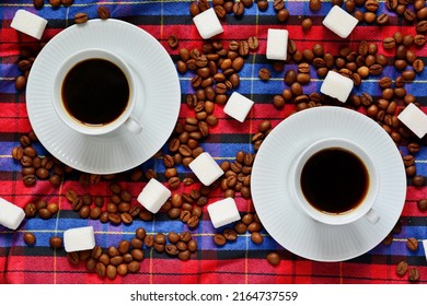 Couple Of Cups Of Hot Black Coffee On White Saucers Between Scattered Roasted Coffee Beans And Refined Sugar Pieces On Checkered Fabric. Top View, Flat Lay