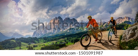 Couple cycling on electric bike, rides mountain trail. Woman and Man riding on bikes in Dolomites mountains landscape. Cycling e-mtb enduro trail track. Outdoor sport activity.