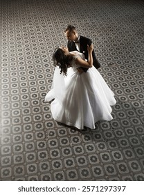 couple dancing in a large room with a patterned floor. The woman is wearing a white dress and the man is wearing a dark suit.