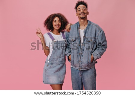Couple in denim outfits posing on pink background. Dark-skinned woman and brunette man in white tees smiling on isolated