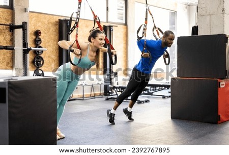 A couple of different ethnicities perform some exercises with special tapes inside a gym. Young people work their pectorals using suspension straps. Concept of suspension exercises.
