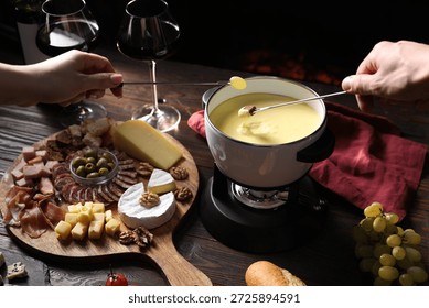 Couple dipping piece of bread and grape into fondue pot with delicious melted cheese at wooden table with snacks against black background, closeup