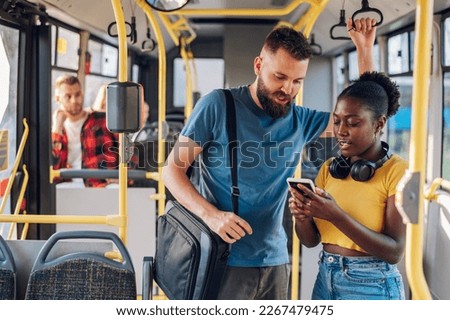 Couple of diverse young friends standing and talking together on a bus and using a smartphone. Multiracial people having fun while commuting by a bus. Public transport, technology and people concept.