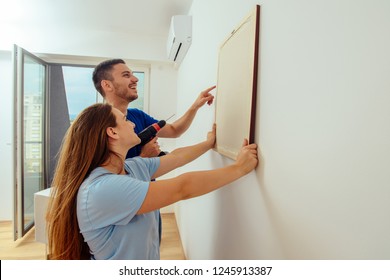 Couple Is Doing Repair And Hanging Picture On The Wall At Home. Young Family Moving In