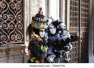 Couple Dressing Carnival Costumes In Venice Italy. Carnival Dresses Made With Black And White Plumes And Coloured Squares Like Gaming Cards. On The Background Handcrafted Iron Gates. Venice, Italy.