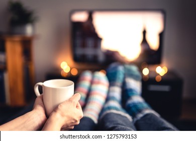 Couple drinking tea, hot chocolate, eggnog or mulled wine and watching tv in warm cozy woolen socks in winter. Woman holding cup of morning coffee in home living room. Sick people with flu or fever.