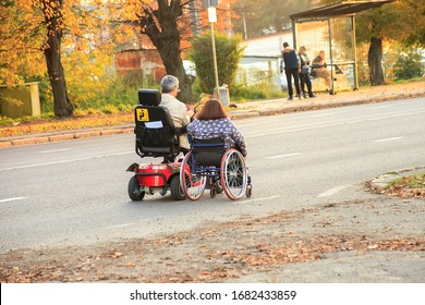 Couple Elderly Disabled People In Electric Wheelchair Driving On Road. Man And Woman On Motorized Wheel Chair Motion