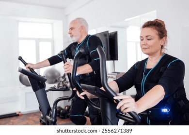 Couple of elderly people in ems suits are engaged in sports equipment. Old man is exercising on elliptical trainer. Aged fat woman pedaling exercise bike. Electrical muscle stimulation training. - Powered by Shutterstock