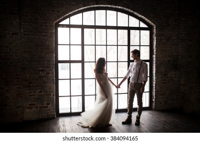 couple embrace on a background of textured walls in the loft, the bride in a wedding dress and beige with a bouquet of anemones in hand, groom in beige trousers and a white shirt with suspenders
