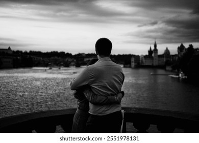 A couple embraces on a bridge, gazing at a calm river and distant historic buildings under a moody sky. - Powered by Shutterstock