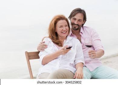 Couple embracing each other's at teh beach. People in love smiling, drinking red wine from glasses and sitting on comfortable chairs.