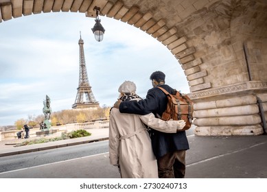 Couple embracing while walking under a stone arch bridge, enjoying a romantic travel sightseeing tour and admiring the iconic eiffel tower on a cloudy day in paris, france