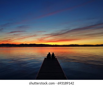 Couple At The End Of A Pier (couple & Pier In Silhouette) Enjoying A Spectacular Orange, Red & Blue Sunset Behind Adirondack Mountains At Lake Champlain In Burlington, Vermont To End A Nice Autumn Day