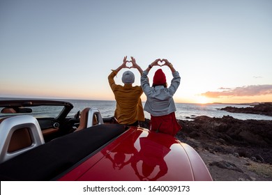 Couple enjoying beautiful views on the ocean, standing together near the car on the rocky coast, showing with hands heart shape. Carefree lifestyle, love and travel concept